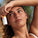 Woman with sun-kissed skin holding Tropic Sun Stick near her forehead, with sand on her arm and a blurred beach background.